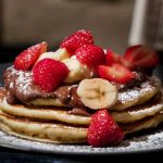 five pancakes sat on a plate with banana and strawberry on top