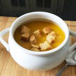 A white bowl full of lentil soup and croutons on a wooden chopping board
