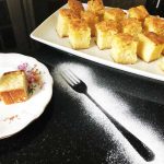 White chocolate and macadamia blondies on a white plate with an icing sugar fork on a black granite worktop