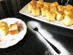 White chocolate and macadamia blondies on a white plate with an icing sugar fork on a black granite worktop