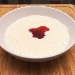 A bowl of stove top rice pudding in a dish with strawberry jam on a bamboo chopping board