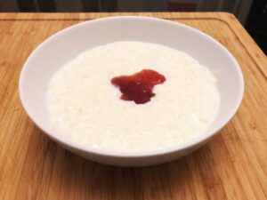 A bowl of stove top rice pudding in a dish with strawberry jam on a bamboo chopping board