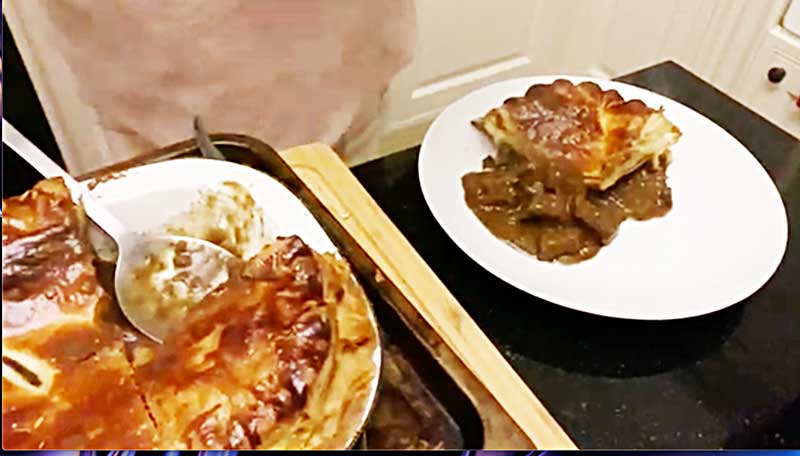 A large steak pie on a wooden chopping board and a white plate with a portion of steak pie on it