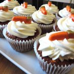 Nine cupcakes on a white plate topped with cream cheese frosting and a chocolate carrot